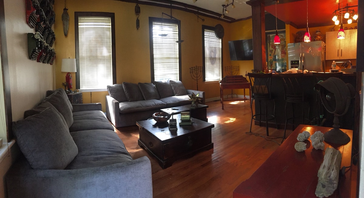 A bright living area features a cozy grey sectional sofa, complemented by a dark wood coffee table at its center. Two windows allow natural light to fill the space, while colorful pendant lights hang above the adjoining kitchen area, visible in the background.