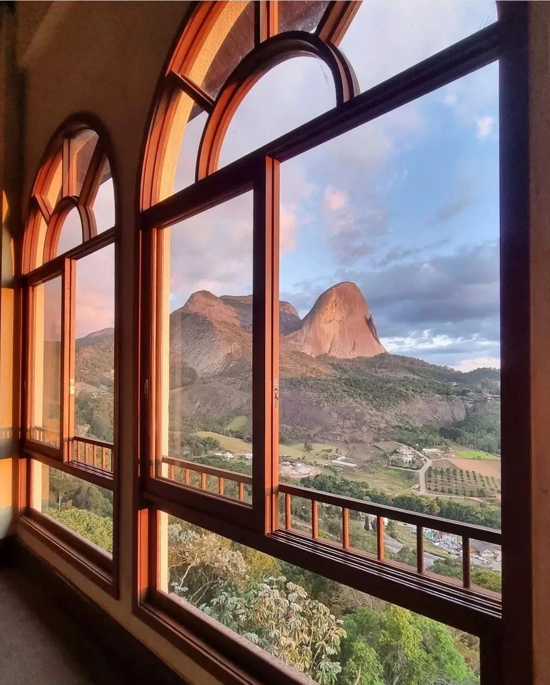 Large arched windows frame a stunning view of Pedra Azul, capturing the mountain's unique silhouette against a colorful sky. The landscape below includes greenery and cultivated fields, providing a serene connection to the outdoors.