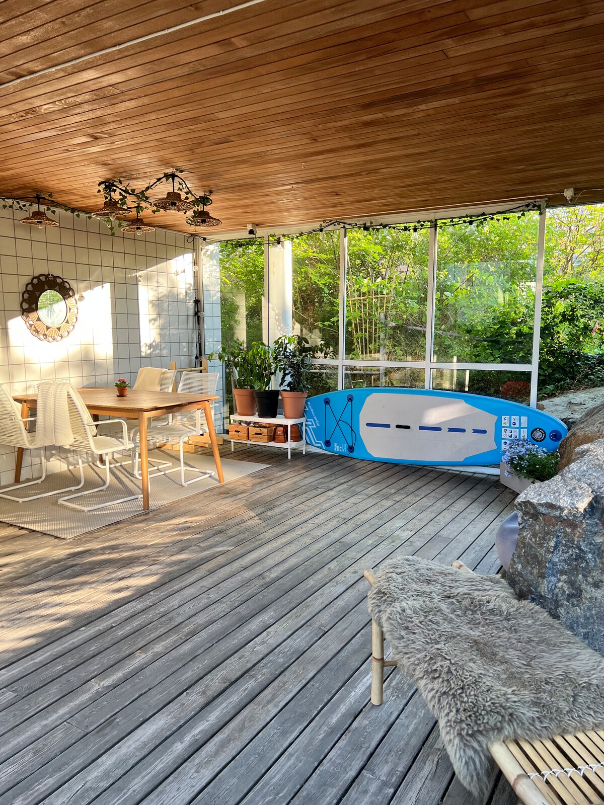 A cozy terrace features a wooden deck and is surrounded by greenery. A dining table with four chairs is positioned beside decorative plants. A large window allows natural light to fill the space, while a surfboard rests against the wall, adding a casual touch.