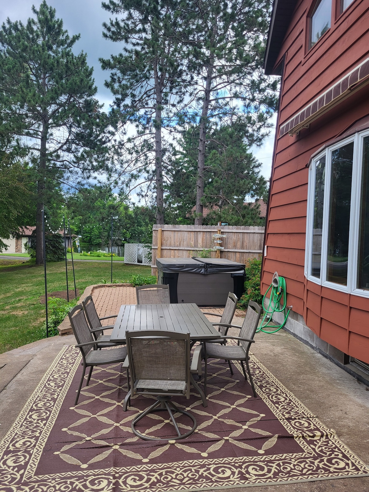 A patio area is shown with a rectangular table surrounded by six coordinated chairs. A decorative outdoor rug is placed beneath the table, adding a touch of style. Surrounding trees and greenery contribute to the natural ambiance, while a grill and garden hose are visible nearby.
