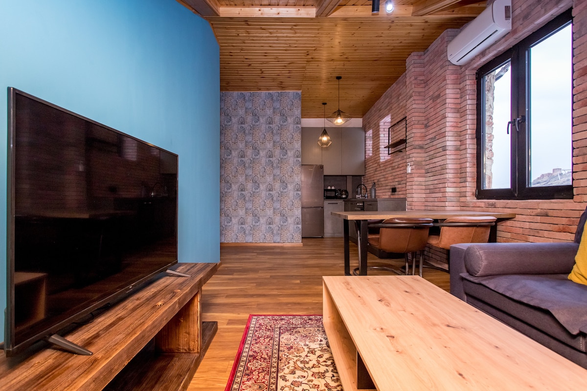 A cozy living area features a wooden coffee table and a comfortable sofa, complemented by a large television on a rustic shelf. The backdrop consists of a blue accent wall and exposed brick, with a well-equipped kitchen visible in the distance. Natural light enters through the large windows.