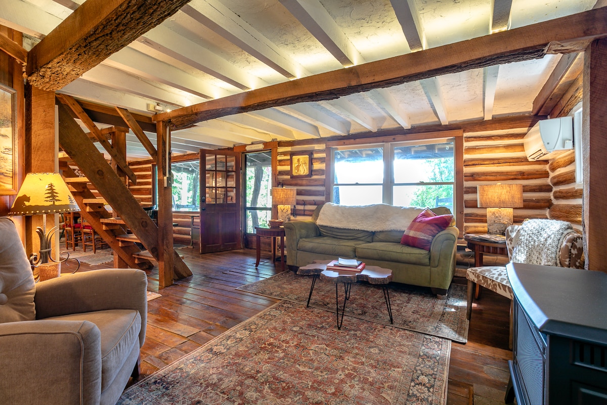 The open living area features warm wooden beams and a rich wood floor, creating a rustic atmosphere. A comfortable couch and armchair are arranged for relaxation, while large windows provide light and views of the surrounding nature. A dining area and wooden furniture are visible in the background.