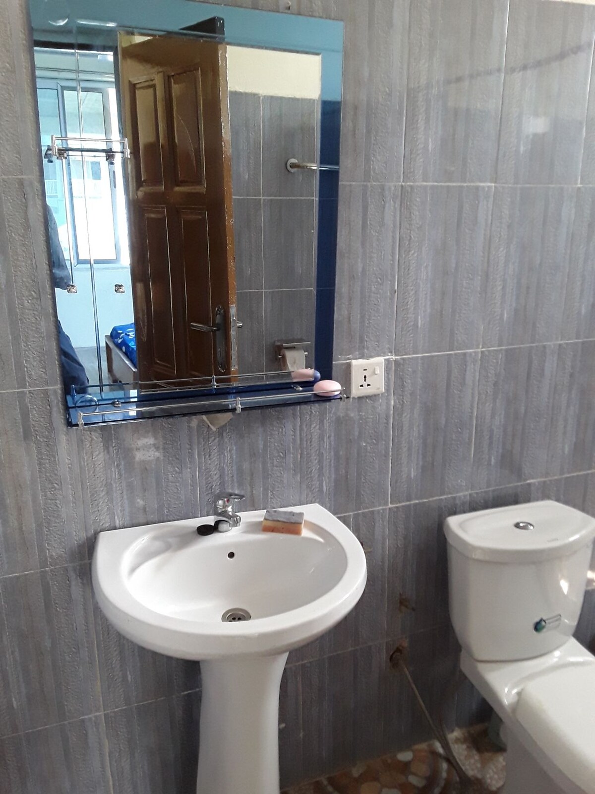 A bathroom features tiled walls in shades of gray. A small white sink is mounted on a pedestal, equipped with a single faucet. A mirror with a blue border is positioned above the sink. A toilet is located adjacent to the sink, completing the restroom setup.