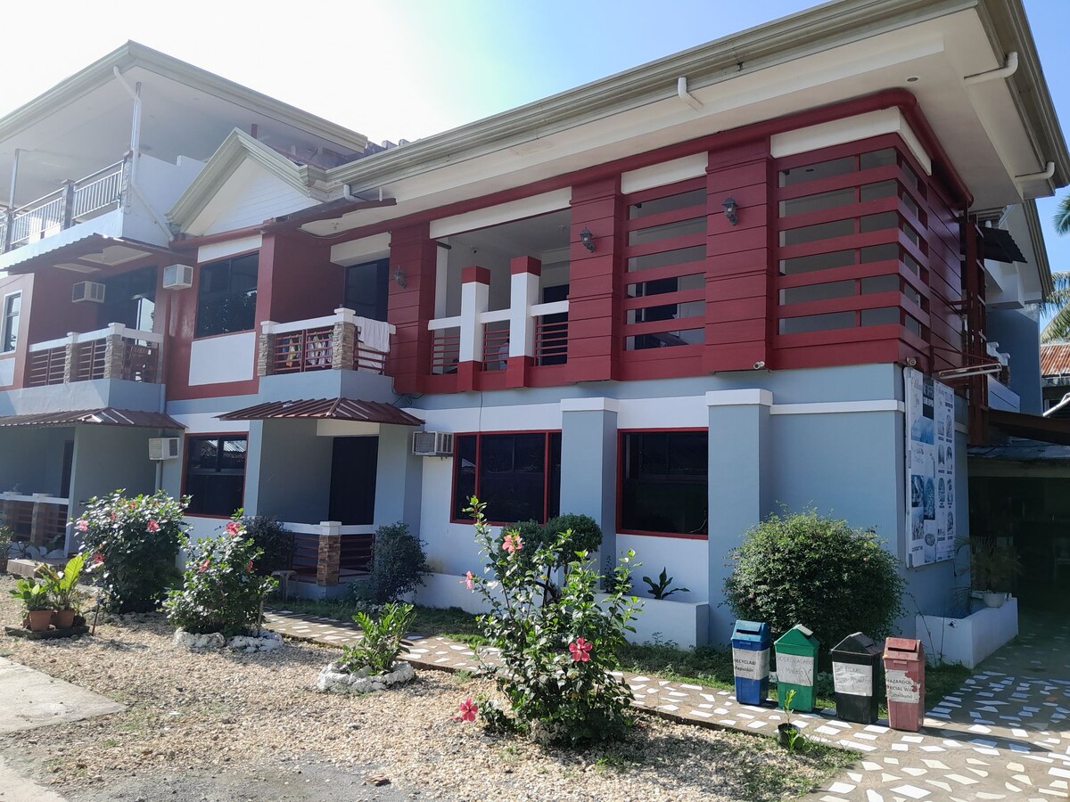 A two-story building is shown, featuring a modern design with red and white accents. Large windows allow natural light to brighten the exterior, complemented by greenery and flowering plants in the foreground. Pathways are lined with decorative stones leading to the entrance.