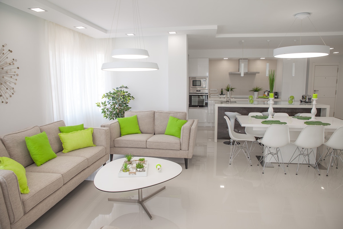 A modern and spacious living area features light-colored furniture accented with green cushions. A round coffee table sits at the center, while a sleek dining area with a white table and chairs is visible in the background. Large windows allow natural light to fill the space.