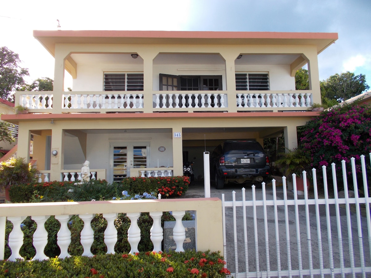 A two-story house is displayed with a white picket fence in front. The ground floor features a covered driveway accommodating a vehicle, with vibrant flowers and landscaping visible. A spacious balcony extends above, adorned with a decorative railing, enhancing the inviting exterior.