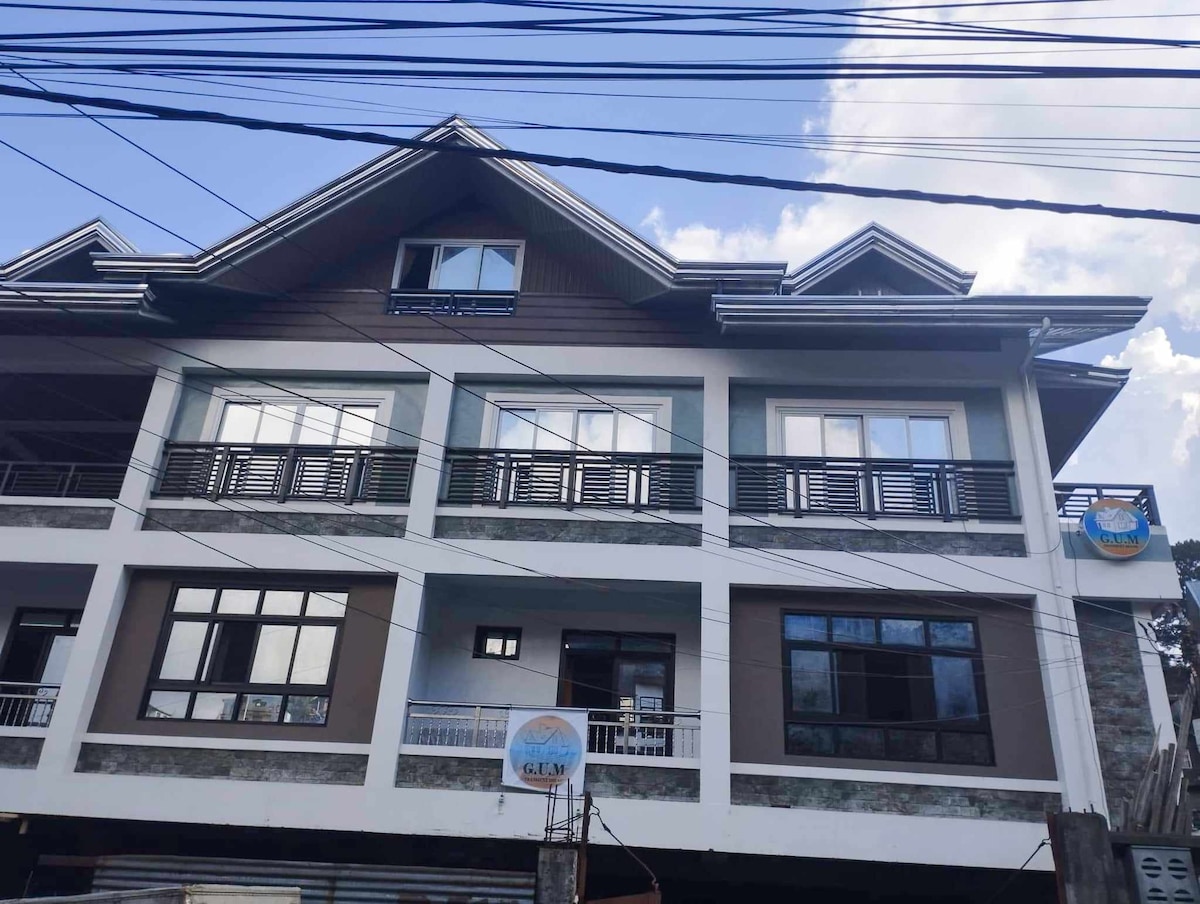 A three-story modern building is depicted with a prominent roof and multiple balconies. The faรงade features a combination of light and dark colors with large windows that provide ample natural light. A sign for G.U.M Transient House is visible above the main entrance.