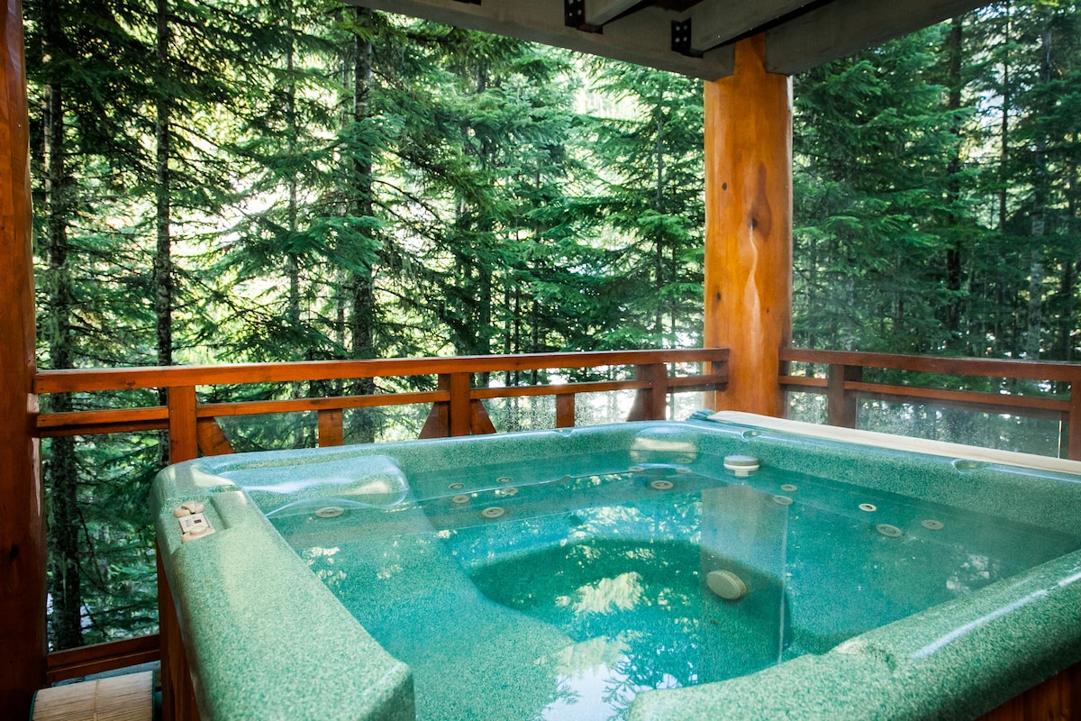 A private hot tub is situated on a deck surrounded by tall evergreen trees, creating a serene outdoor space. The tub features several jets and a textured surface, with wooden railings providing a natural finish that complements the forested backdrop.