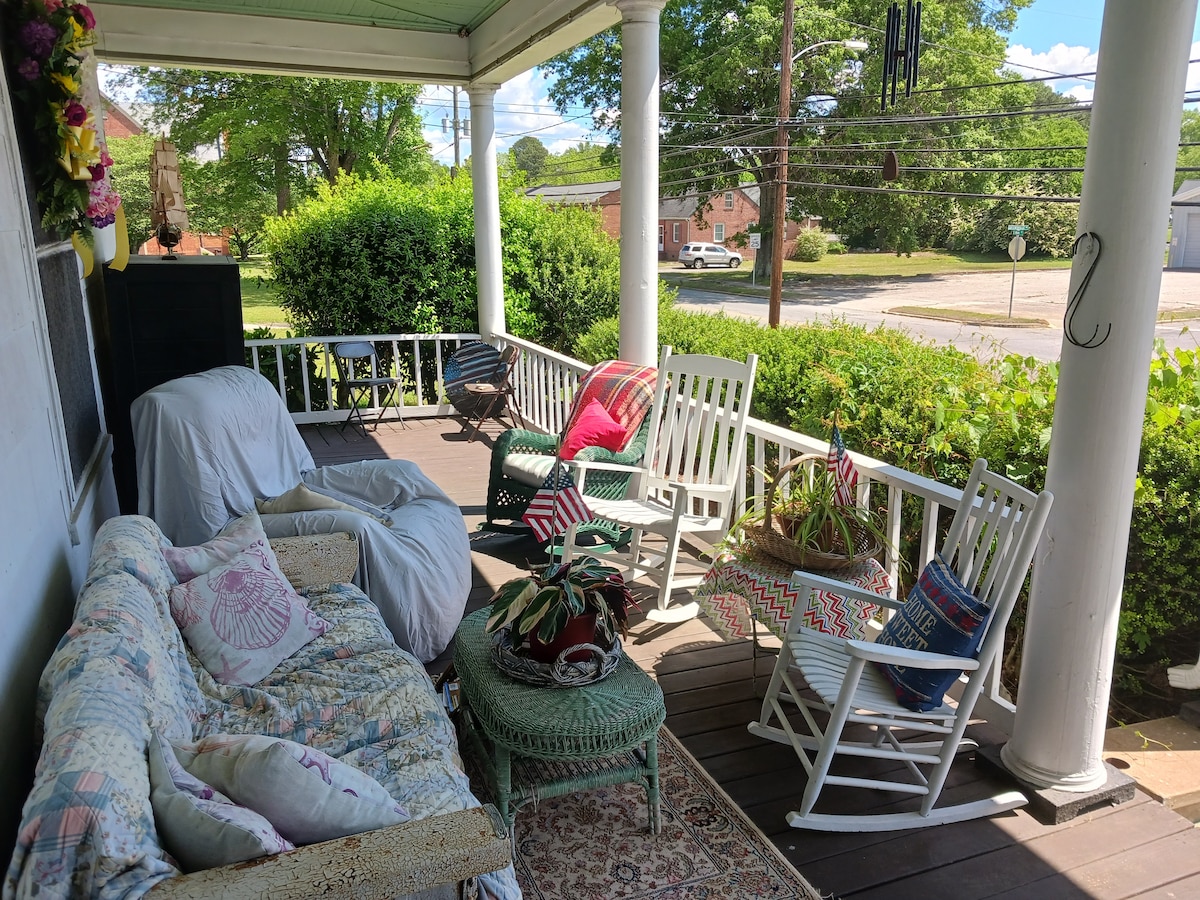 A spacious front porch features a combination of seating options, including rocking chairs and a cozy sofa draped with a quilt. Potted plants and decorative pillows add color, while greenery and a view of the neighborhood create a welcoming atmosphere.