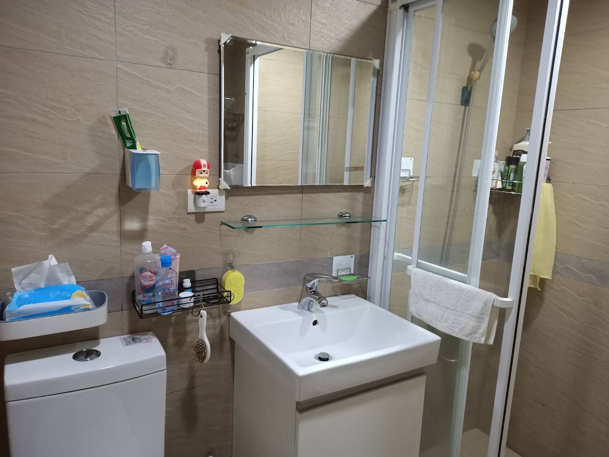 The bathroom features a modern design with a glass shower enclosure and a wall-mounted mirror above the sink. Toiletries are neatly arranged on shelves, and a small shelf holds additional supplies. A towel is hung conveniently near the shower.