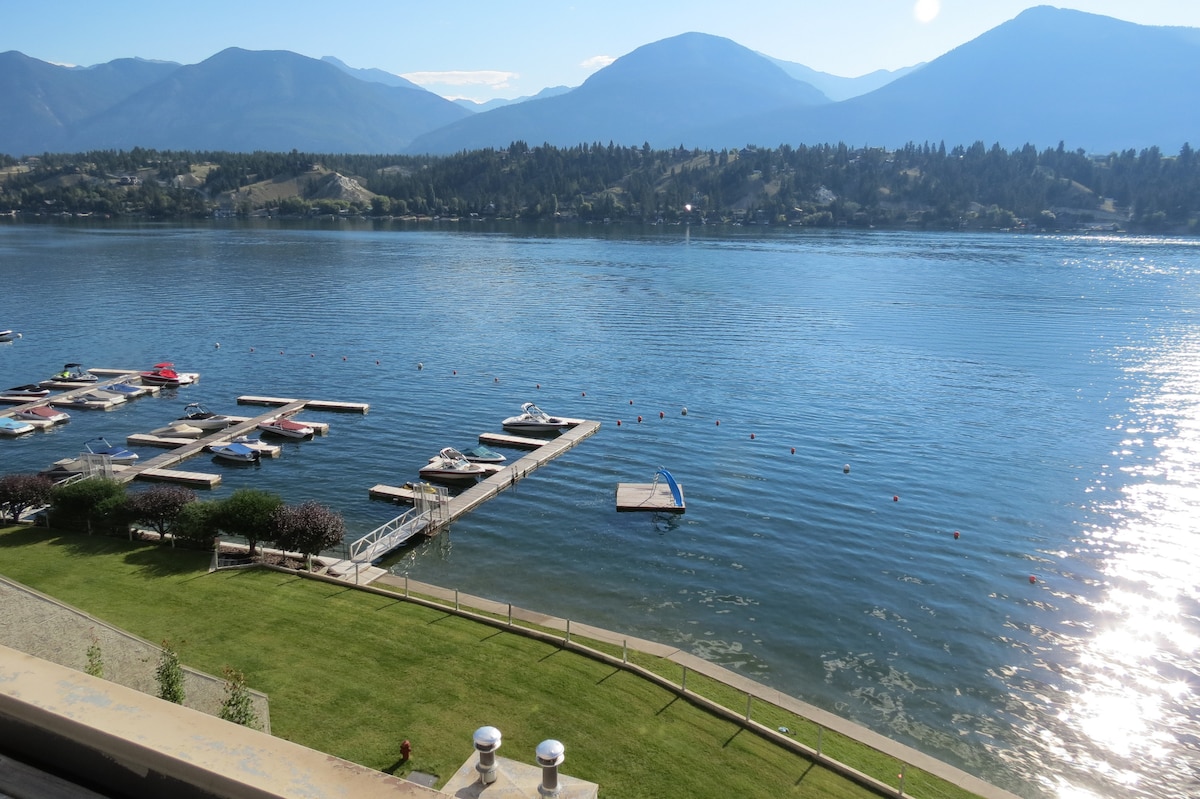 A serene view of the crystal-clear lake is presented, showing several docks with boats anchored. Lush green grass lines the shore, leading to the water's edge. Majestic mountains rise in the background under a clear blue sky, reflecting off the calm lake surface.