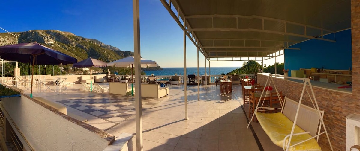 The spacious terrace offers stunning sea views and is equipped with outdoor seating and shaded areas. Comfortable lounge chairs and swings are visible, providing space for relaxation. The flooring features a distinct pattern, enhancing the open atmosphere of the area.