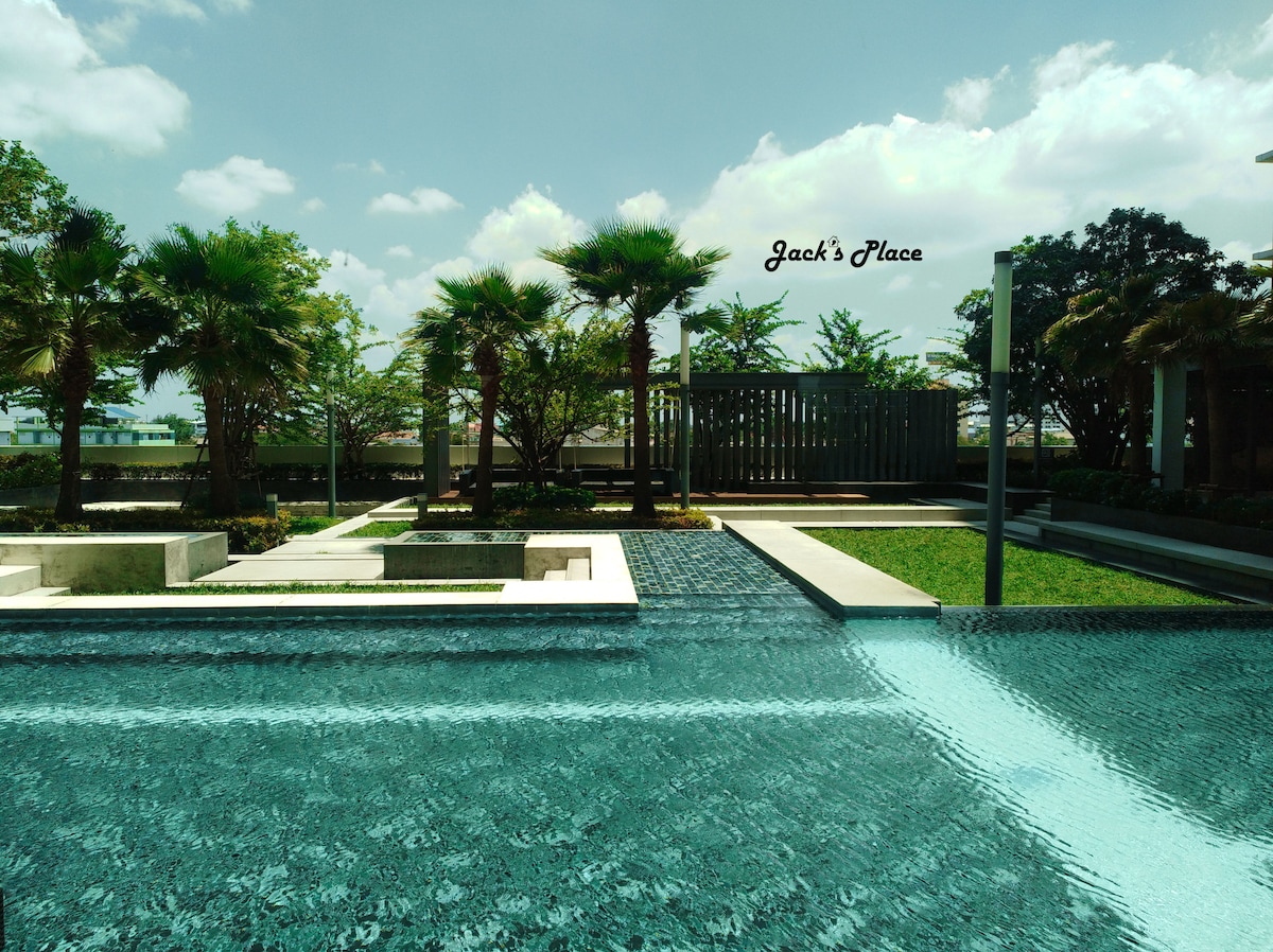 A serene outdoor pool area features a clear water surface reflecting the sky above. Lush palm trees and manicured greenery provide a natural backdrop, while the surrounding space includes stone pathways and seating areas, promoting a tranquil environment for relaxation.