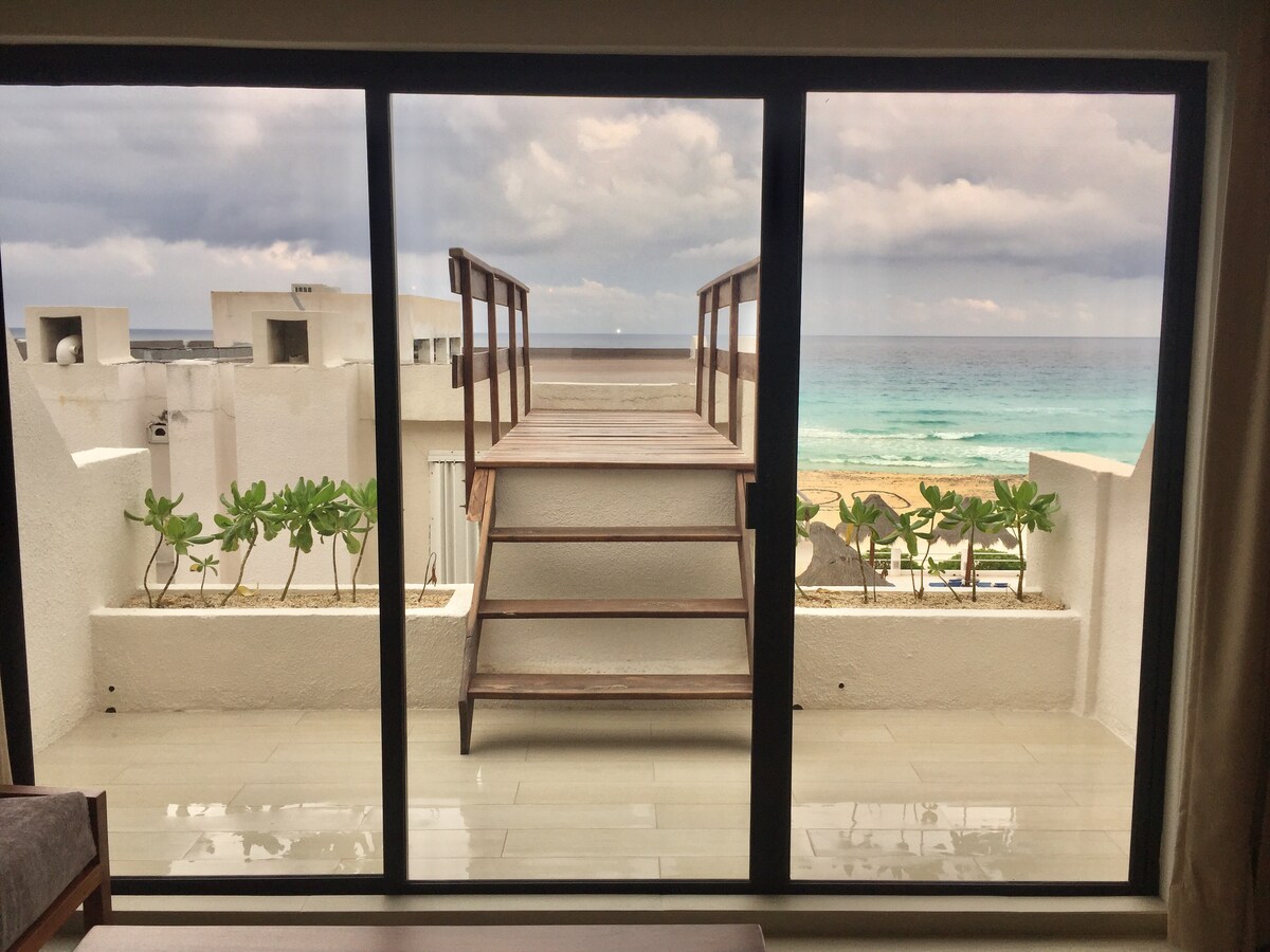 A glass door frames a scenic view of the ocean, featuring a wooden staircase leading to an outdoor deck. Lush greenery is planted along the terrace edge, contrasting with the soft blues of the sea and the cloudy sky.