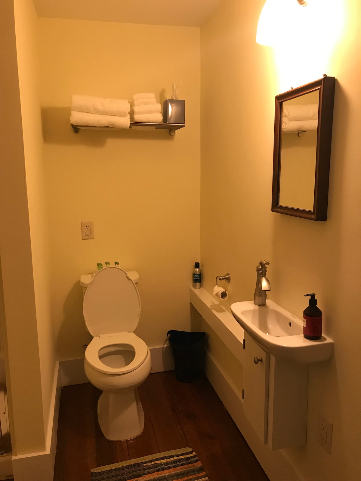 A well-appointed bathroom features a white toilet and a compact sink with a modern faucet. Fresh towels are neatly arranged on a shelf above. A small mirror hangs above the sink, complemented by a wall light providing illumination. A colorful rug adds a touch of warmth to the wooden floor.