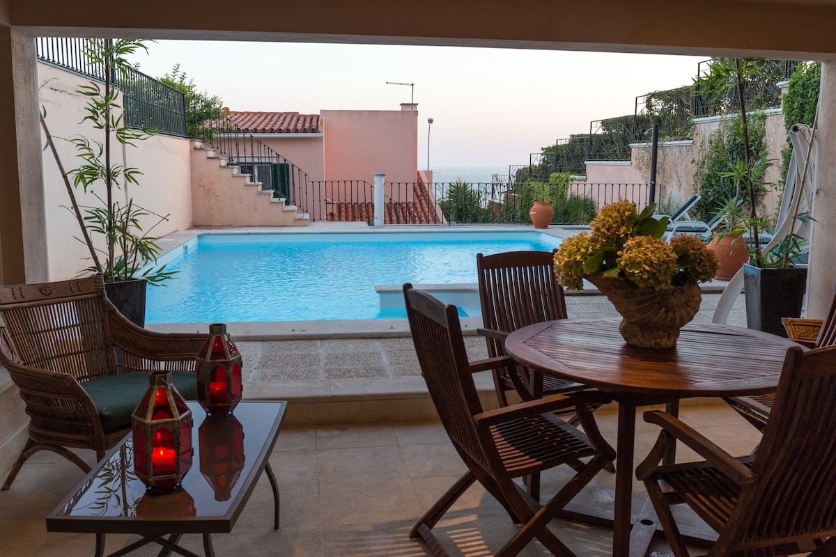 An outdoor seating area is presented with a round wooden table and four wooden chairs. Nearby, a vase of flowers adds a touch of nature. A refreshing salt-water pool is visible in the background, framed by lush greenery and tranquil pathways.