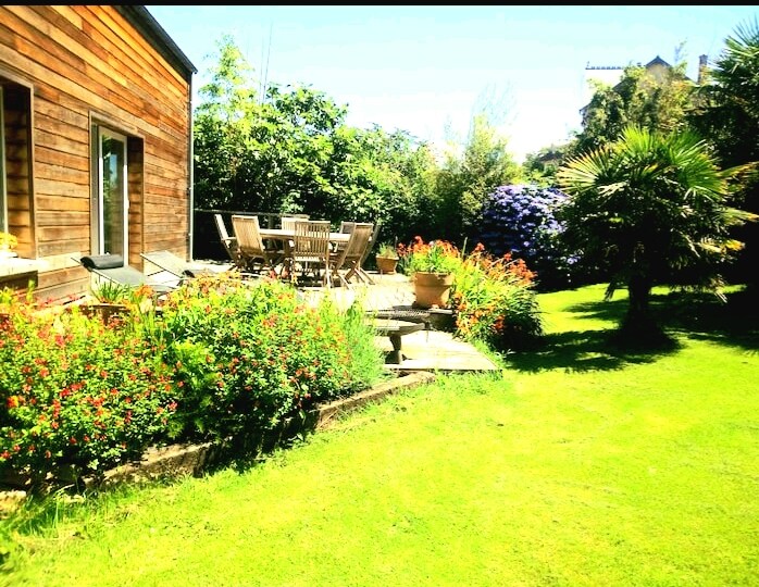 A garden area features a well-maintained lawn framed by colorful flowers and greenery. A wooden patio set with multiple chairs surrounds a table, providing a space for outdoor dining. Sunlight enhances the vibrant colors of the plants and the natural wood of the building.
