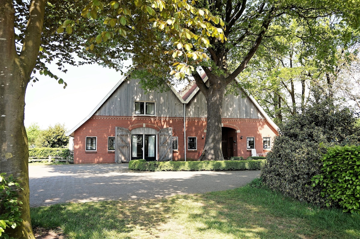 A charming farmhouse is set amidst greenery, featuring distinctive architectural elements. Large open doors reveal the inviting entryway, while surrounding trees provide shade and enhance the serene atmosphere. A well-maintained lawn borders the property, contributing to the overall pastoral setting.