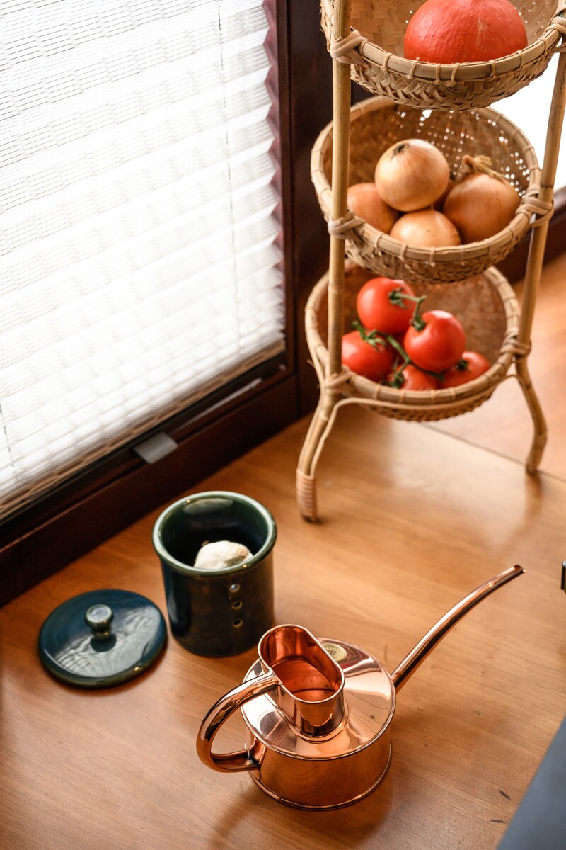 A copper watering can is positioned beside a ceramic container with a lid. A woven basket, placed on a wooden surface, showcases ripe tomatoes and apples, enhancing the kitchen's functional decor. Natural light filters through a textured window covering, providing a warm ambiance.