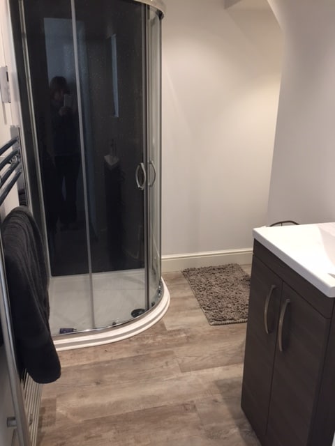 A modern bathroom is presented with a glass shower enclosure and a sleek vanity unit. Light-colored walls create a clean atmosphere, complemented by a textured mat on the floor. A towel hangs from a rack, adding functionality to the space.