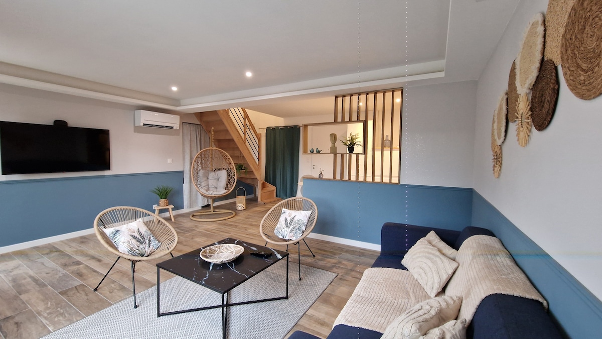 A spacious living area features a combination of light blue and neutral walls, with a comfortable sofa and two accent chairs. A stylish coffee table centers the space, while a large television is mounted on the wall. A staircase leads to the upper level, adding depth to the room.