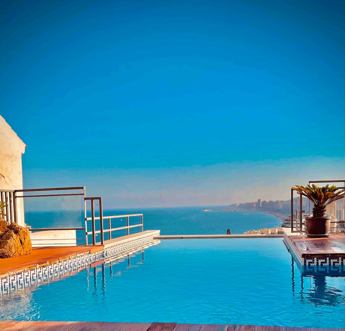 A tranquil infinity pool is visible, merging with the horizon and ocean beyond. Clear blue skies are reflected on the water's surface, while a terracotta planter with greenery is situated nearby. A wooden deck surrounds the pool, enhancing the serene seaside atmosphere.