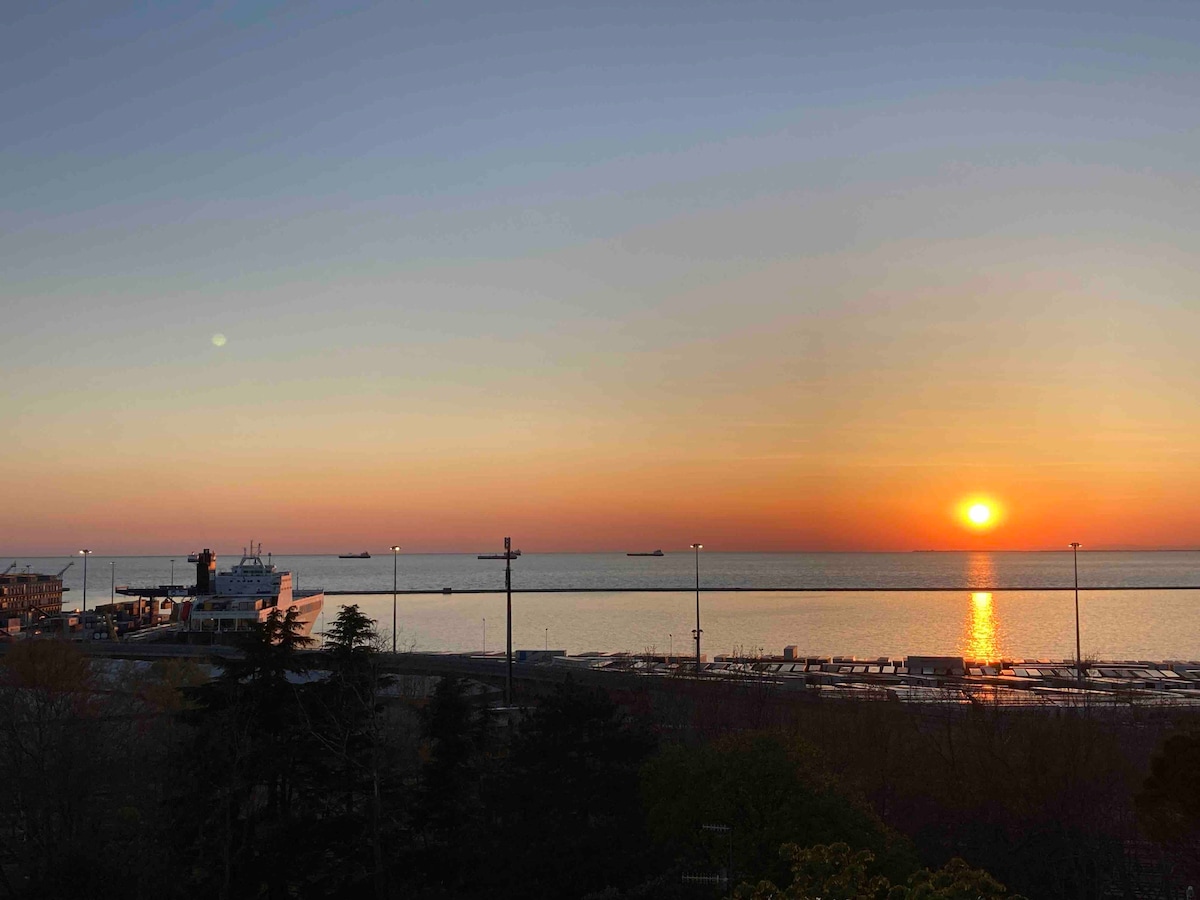 An expansive view of the sun setting over the calm waters of the gulf, casting warm orange hues across the sky. Silhouettes of distant ships and shoreline structures are visible against the soft gradient of colors.
