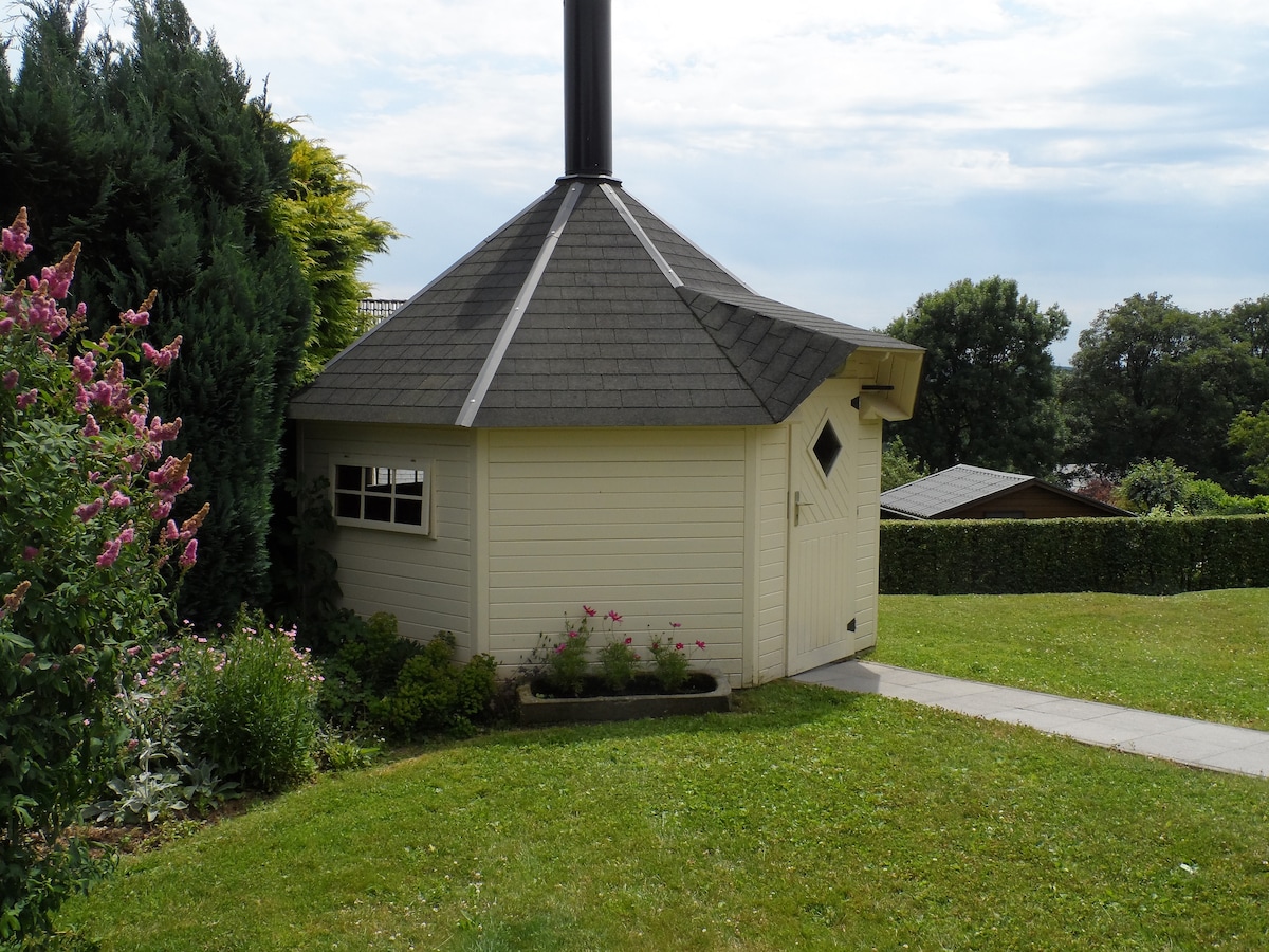 A charming barbecue cabin is nestled within a well-maintained garden, framed by blooming flowers. The octagonal structure features a distinctive chimney and a single door, complementing the surrounding greenery and open lawn.