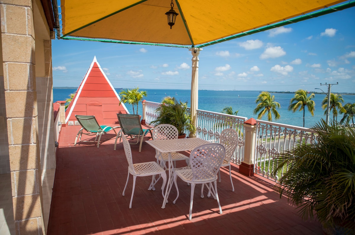 A spacious terrace features a round table with four white chairs, shaded by a large yellow awning. Two lounge chairs are positioned alongside, offering views of the water and palm trees under a clear blue sky.