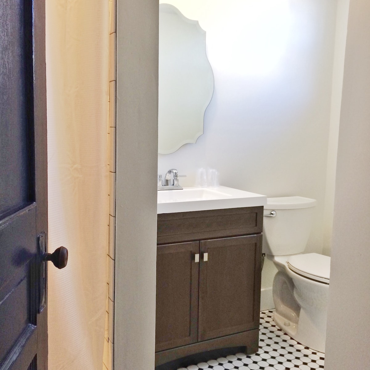 A private bathroom is showcased, featuring a modern vanity with a white countertop and storage cabinetry. A toilet is positioned beside the vanity. The floor is adorned with black and white patterned tiles, and a decorative mirror is mounted on the wall.