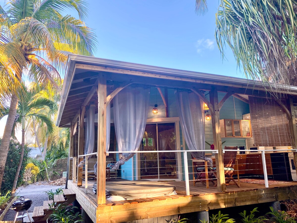 A wooden bungalow is surrounded by palm trees, showcasing a covered terrace with sheer curtains. The entry is marked by a wooden deck, and an outdoor dining area is positioned in front. Nearby, a barbecue is visible, contributing to the inviting outdoor atmosphere.