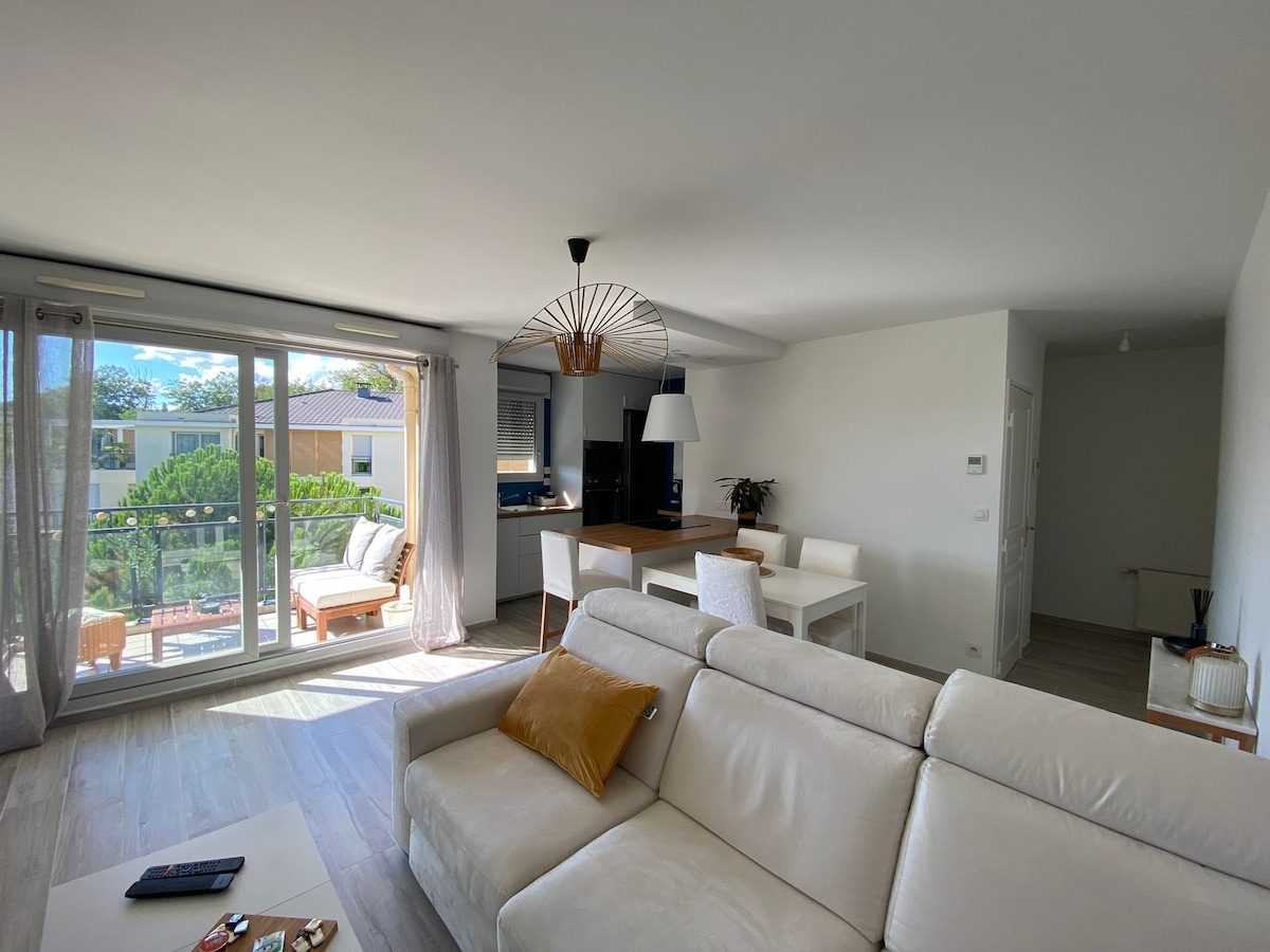 A modern living space features a light-colored sofa adorned with a decorative cushion. A dining area with a white table and chairs is visible. Large glass doors lead to a balcony, allowing natural light to fill the room, enhancing the calming atmosphere.