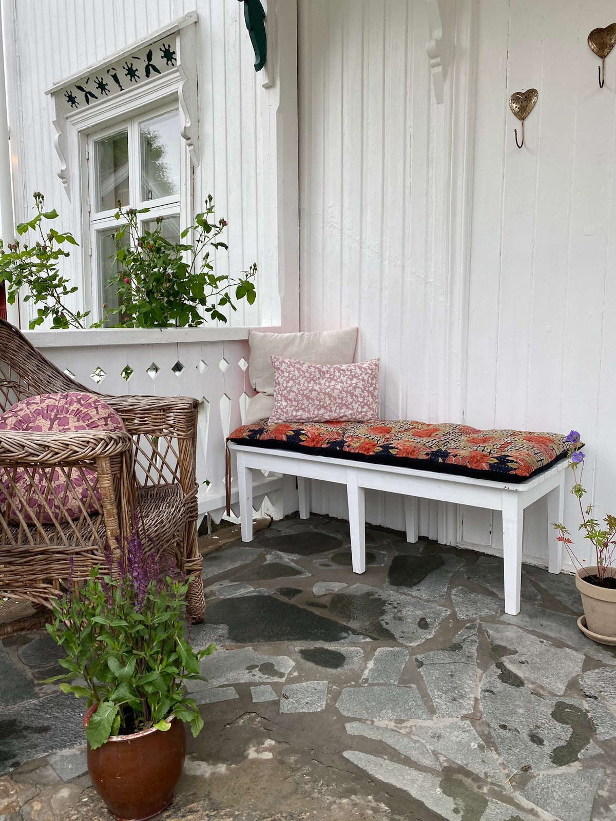 A quaint outdoor seating area is shown, featuring a white bench with a floral cushion against a backdrop of white wooden panels. A wicker chair with decorative pillows is positioned nearby. Potted plants add a touch of greenery, complementing the stone floor.
