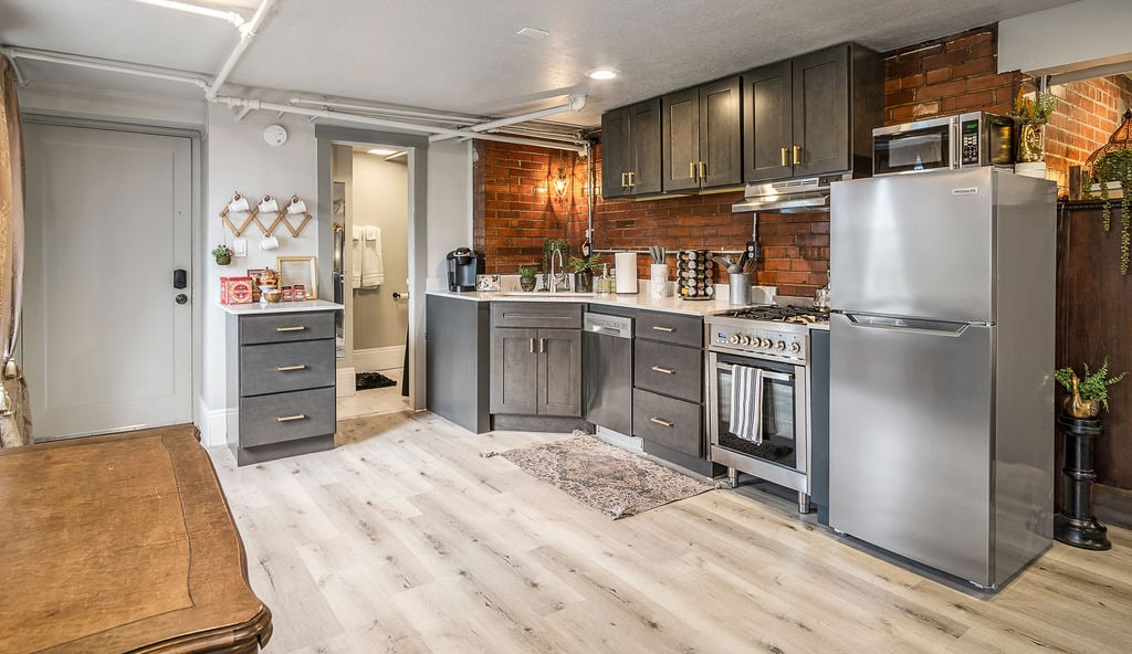 A modern kitchen features dark cabinetry and a contrasting white countertop, with stainless steel appliances including a refrigerator and a gas range. Brick walls add texture, while warm lighting enhances the inviting atmosphere. An area rug lies on the light wooden floor, completing the space.