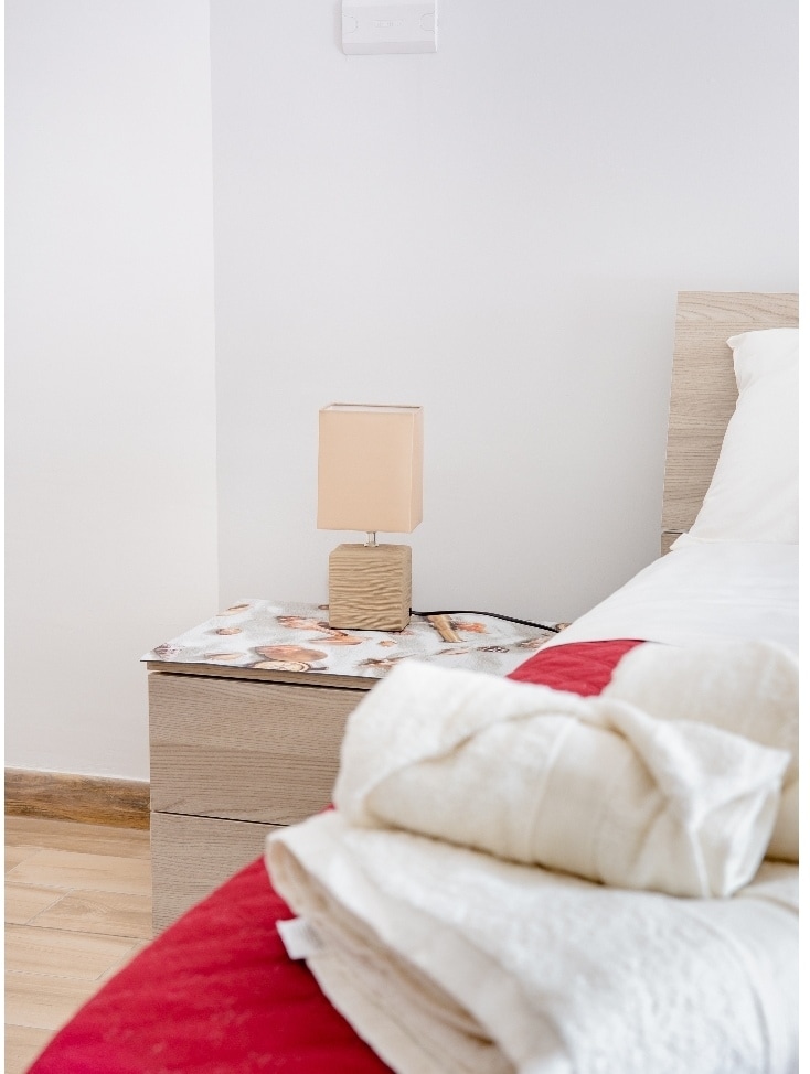 A sleeping area is highlighted, featuring a neatly made spacious bed with red accents. A light wood bedside table is present, topped with a neutral-tone lamp. Soft, folded towels are arranged at the foot of the bed, creating a restful environment.