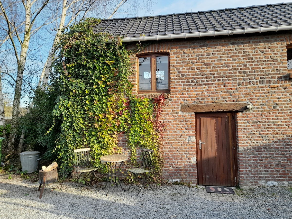 A charming exterior of a brick micro home is displayed, featuring a door with rustic wood accents. Two small chairs and a round table sit on gravel, bordered by vibrant greenery climbing the walls and a tree in the background.