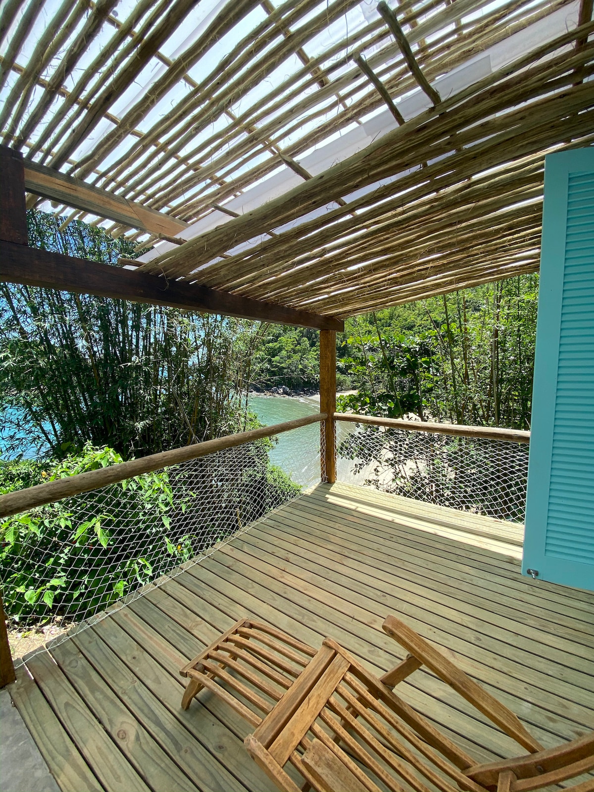 A private balcony with wooden flooring is presented, featuring a simple wooden chair for relaxation. The space is partially shaded by a bamboo roof, surrounded by lush greenery, with views of the nearby beach visible beyond the railing.