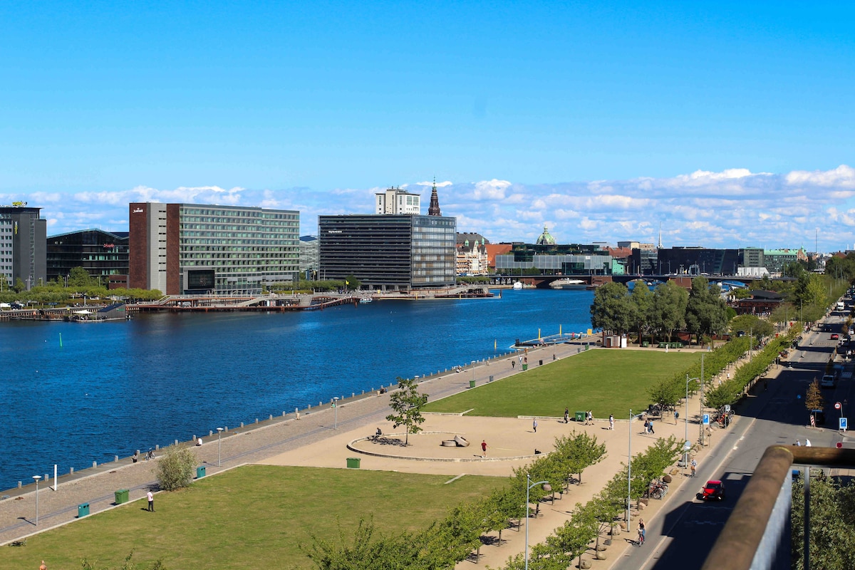 A panoramic view showcases a tranquil waterfront scene, with a serene river lined by greenery. Buildings characterized by modern architecture reflect along the water's surface. People can be seen enjoying the park, adding to the vibrant atmosphere of this waterfront area.