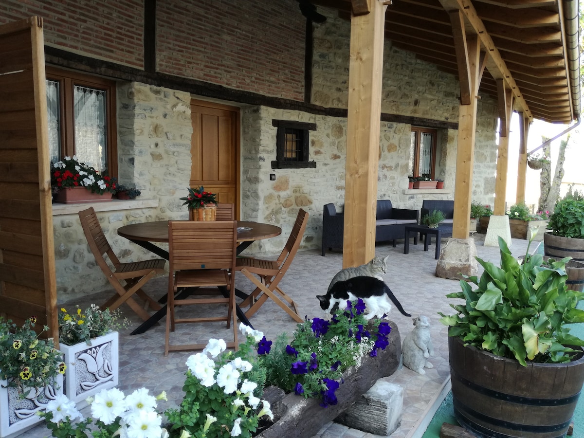 An outdoor porch area is presented with a wooden table surrounded by matching chairs. Comfortable seating is located nearby. Flower pots and planters, filled with vibrant plants, enhance the space, while natural wood beams support the overhang above.