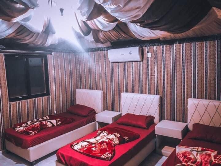 A sleeping area features three spacious beds, each adorned with warm blankets. The walls display a patterned design, complemented by a softly draped ceiling. Large windows allow natural light to filter in, while an air conditioning unit provides climate control.