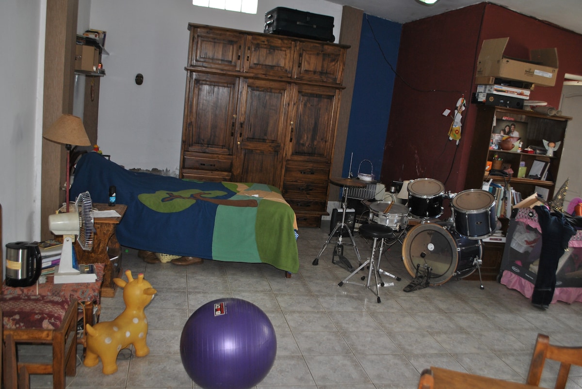 A versatile living area features a cozy sleeping space with a colorful blanket draped over the bed. There is a wooden wardrobe and a bookshelf, along with seating options including wooden chairs. A ball and musical instruments add playful elements to the room.