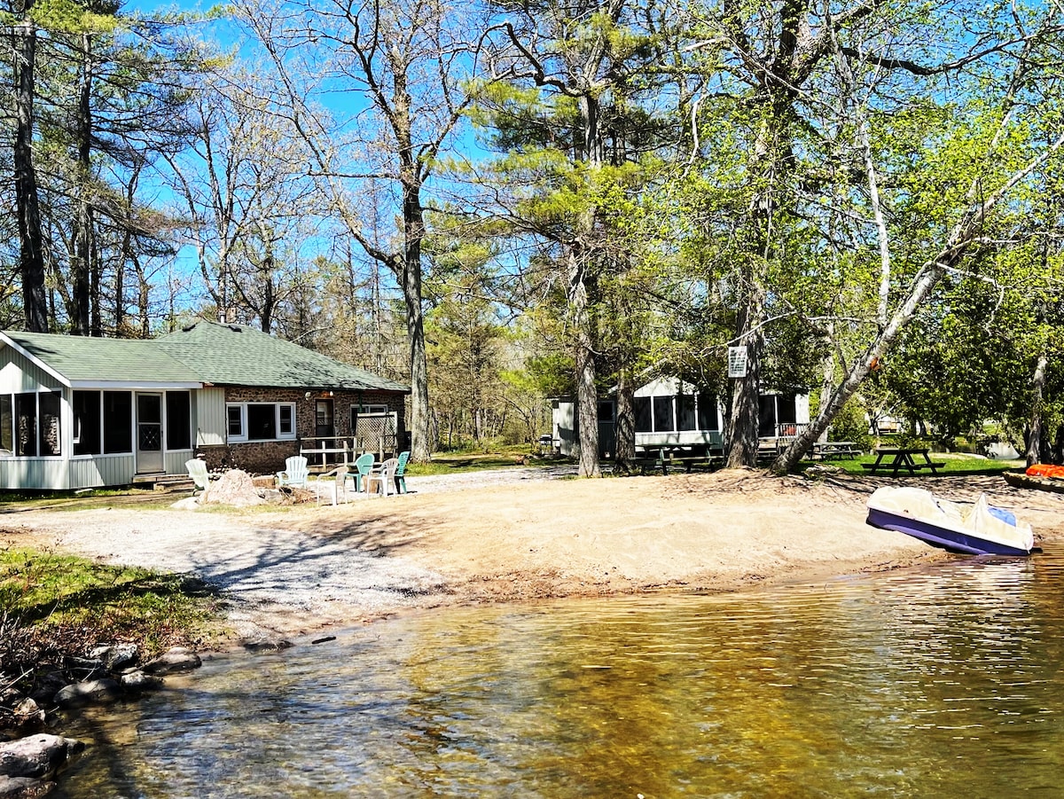 A rustic cottage is visible from the water, with a spacious beach area leading up to it. There are lounge chairs set up near the shore, surrounded by trees. Kayaks and a picnic area are nearby, providing options for leisure activities.