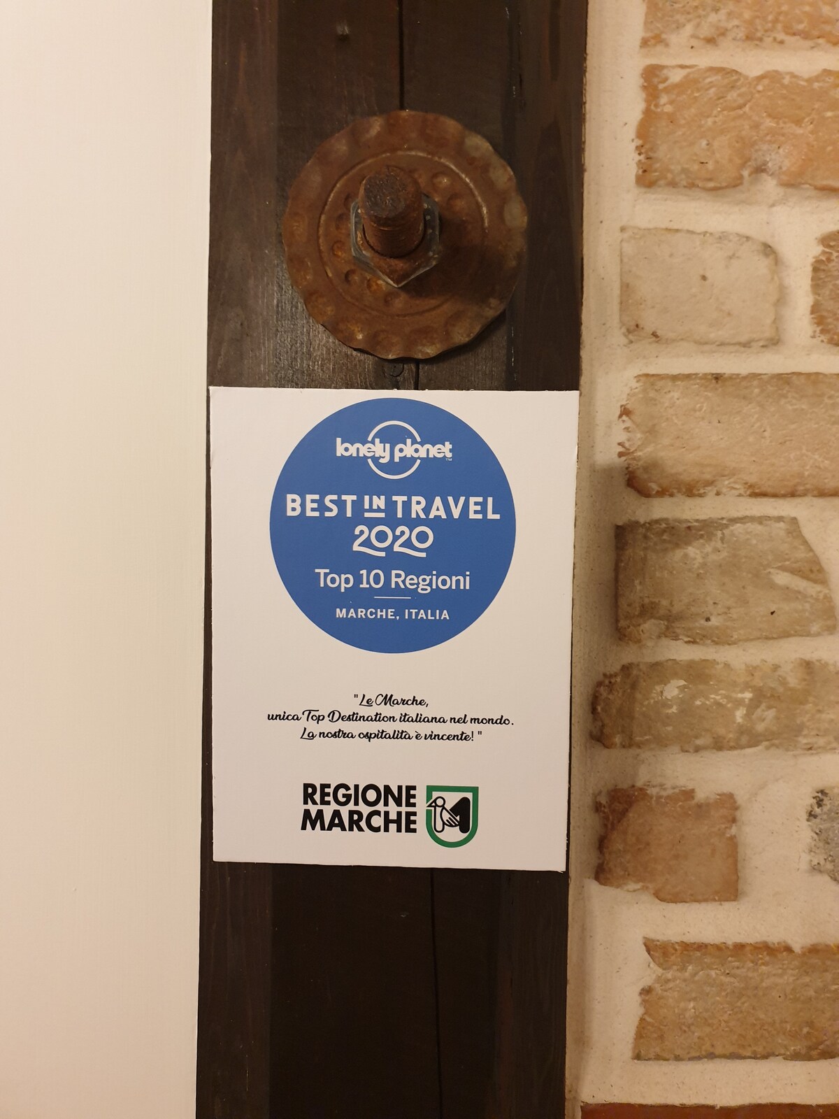 A sign is affixed to a wooden door frame, featuring the Lonely Planet logo. It announces that Marche, Italy, is recognized as one of the Top 10 Regions in the Best in Travel 2020. The door frame is complemented by exposed brick, adding rustic character.