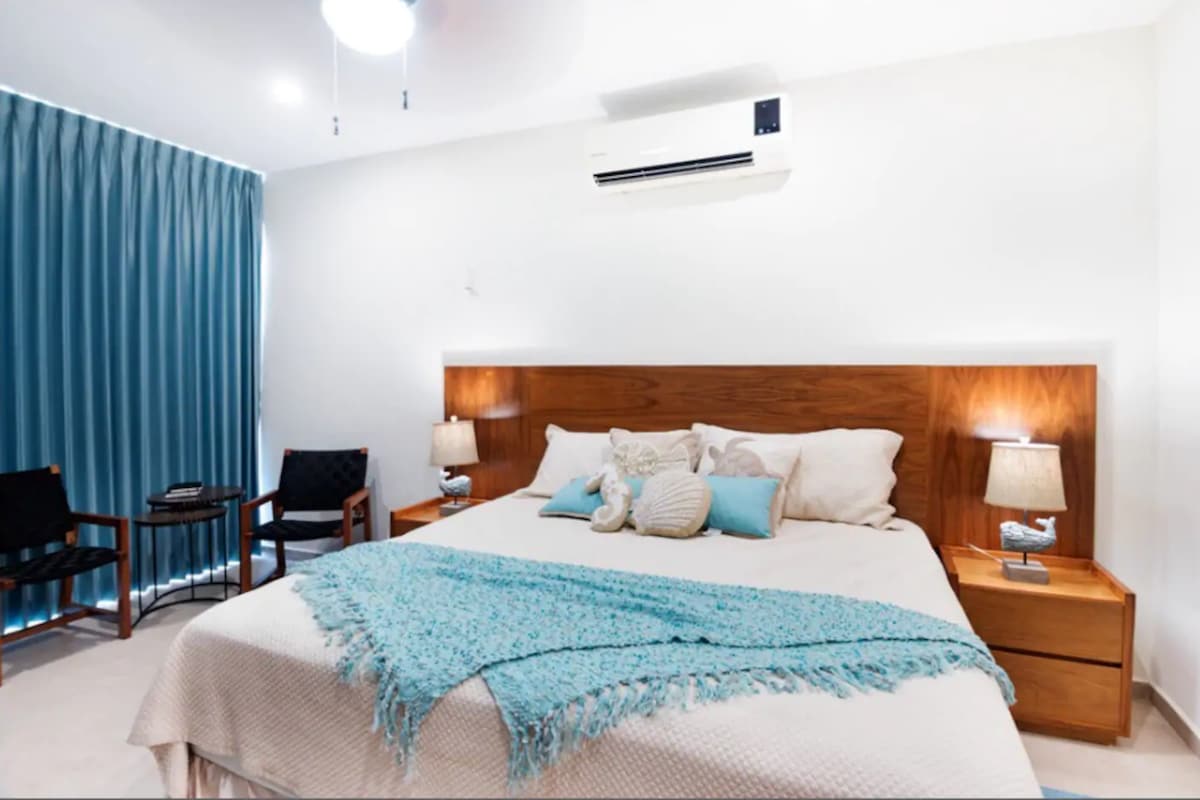 The spacious bedroom features a large bed adorned with soft linens and a textured blanket. Accent lamps provide gentle illumination on either side of the bed. Two stylish chairs are positioned near the window, which is dressed with textured blue curtains.