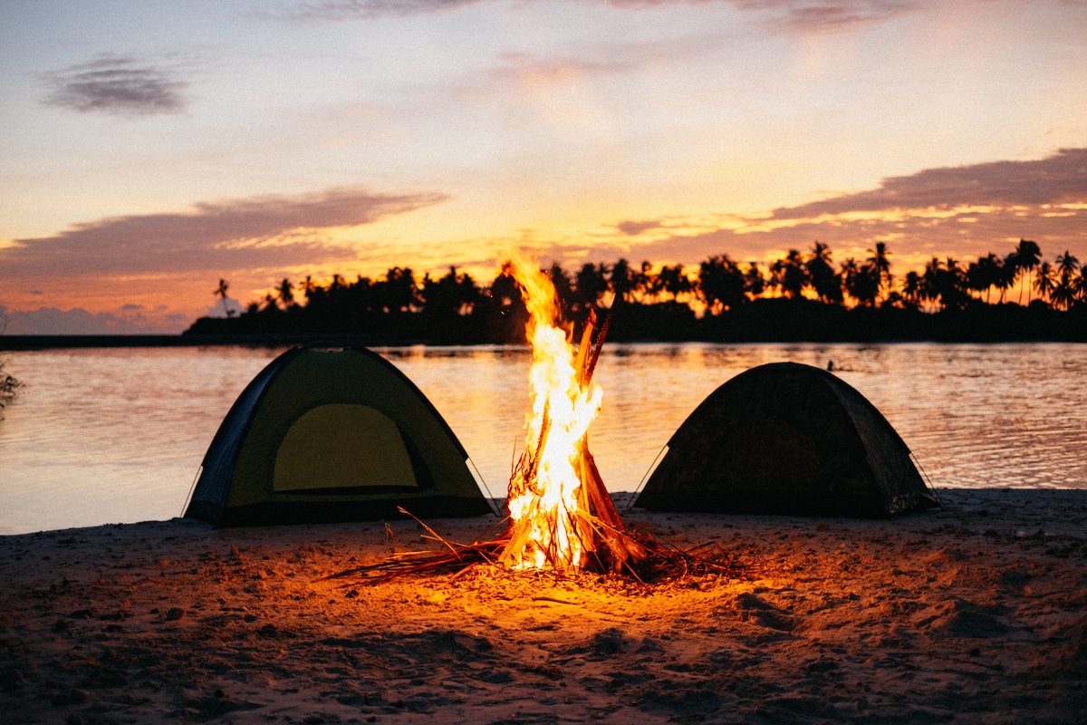 Two camping tents are positioned on a sandy beach, with a crackling campfire between them. The warm glow of the fire illuminates the area, contrasting with the serene sunset over the calm water and silhouettes of palm trees in the background.