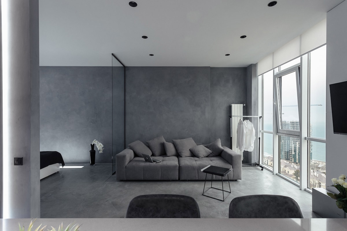 A spacious living area features minimalist grey walls and a comfortable sofa with neutral cushions. A small black coffee table sits nearby, while large windows provide natural light and a view of the sea. A clothing rack is positioned next to the sofa, adding functionality.