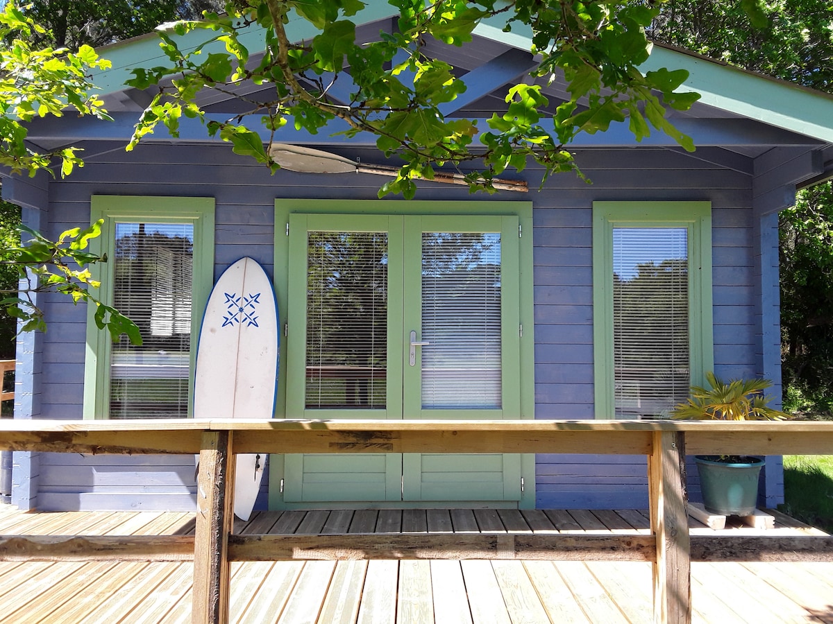 The front of the chalet features a wooden deck with a light-colored railing. Large glass doors are framed by green accents, providing views into the interior. A surfboard leans against the building, while lush greenery surrounds the entrance, contributing to a serene outdoor setting.