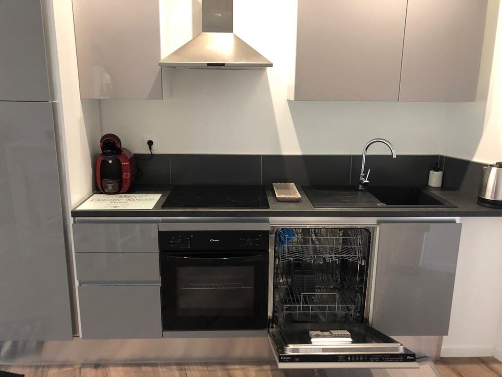 A modern kitchen is visible, featuring sleek grey cabinetry and a black countertop. An open dishwasher reveals space for dishes. A stovetop and a hood are mounted above it, while a coffee maker and kettle are positioned on the counter.