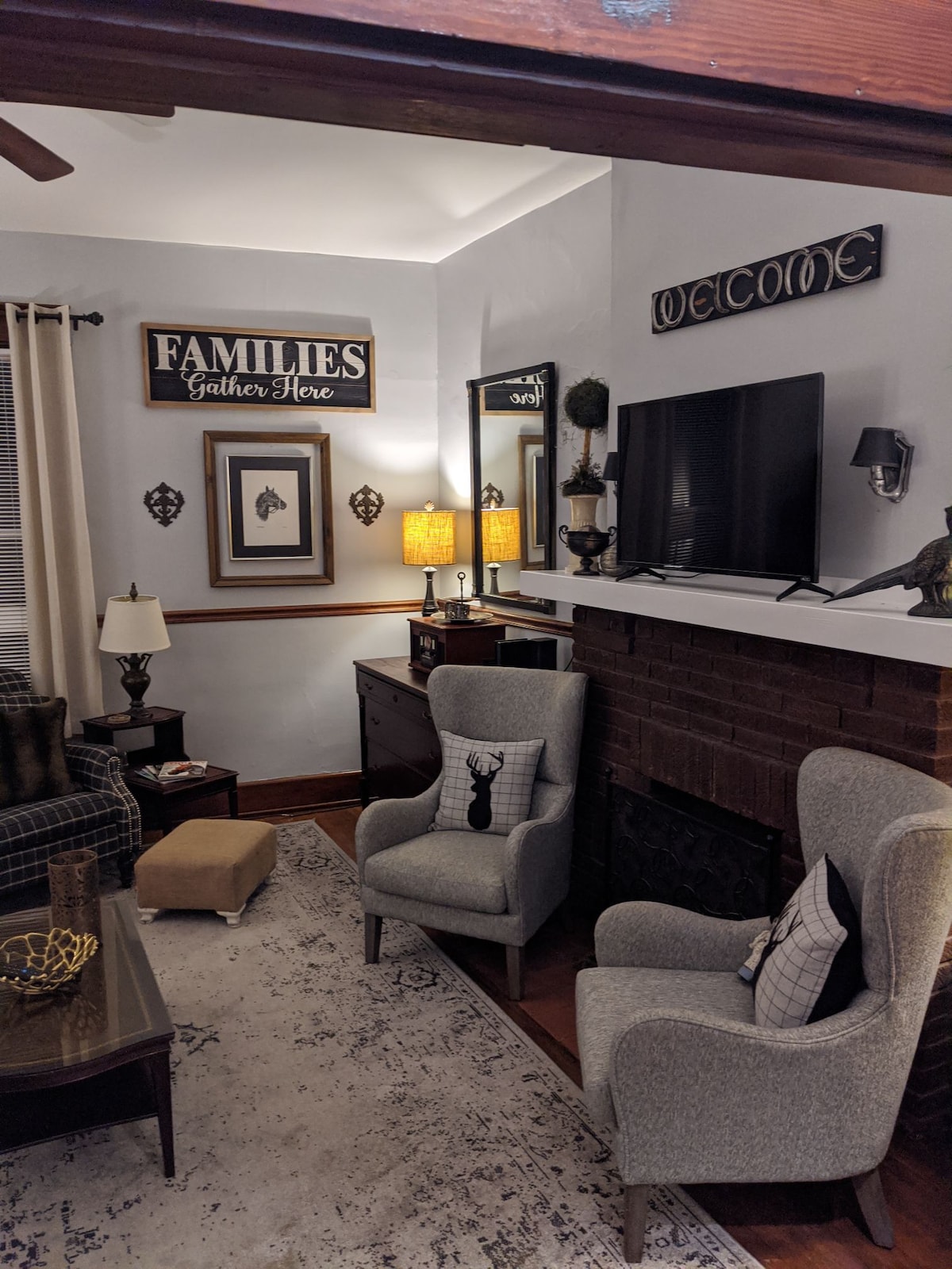 A cozy living room features two light gray armchairs facing a brick fireplace. A modern television is mounted above the mantel, complemented by framed artwork and decorative signs. A warm lamp casts a soft glow, and a patterned area rug anchors the space.