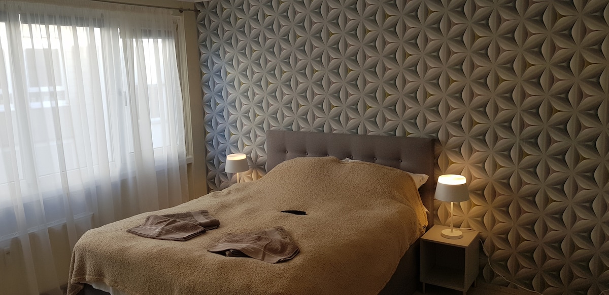 A spacious sleeping area is highlighted by a textured wall covered in a three-dimensional pattern. A soft, neutral bedspread covers the large bed, flanked by two bedside tables with lamps. Natural light filters through the sheer curtains of the expansive window.